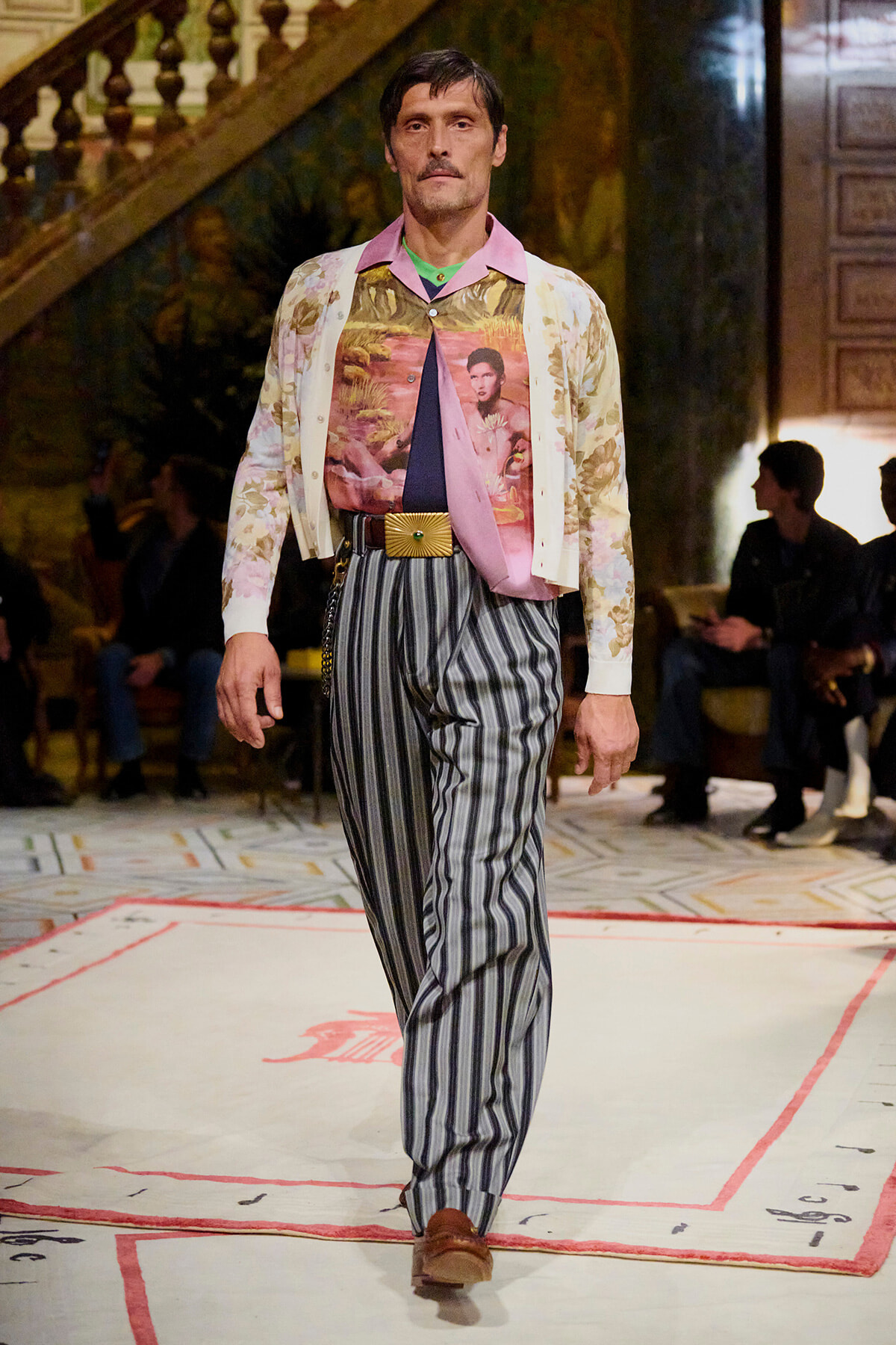 Male model walks a fashion runway in a retro outfit: floral cardigan over a tropical print shirt, striped trousers, and a wide gold belt.