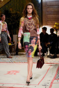Model walking a runway in a color-blocked print dress with face portraits, brown blouse, and burgundy handbag.