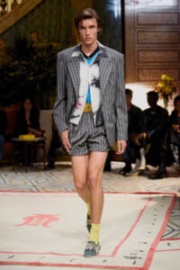 Male model walks the runway in a gray pinstripe blazer and matching shorts, blue top, and yellow socks with blue sneakers.