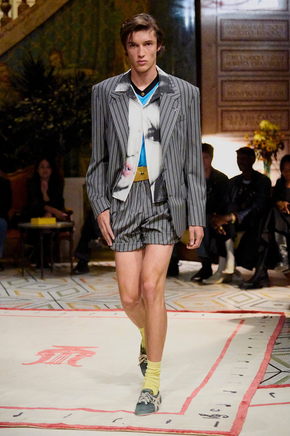 Male model walks the runway in a gray pinstripe blazer and matching shorts, blue top, and yellow socks with blue sneakers.