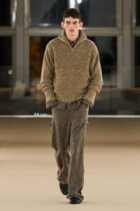 Male model walks a fashion runway in a speckled beige knit sweater and matching loose trousers, hands in pockets, black shoes.