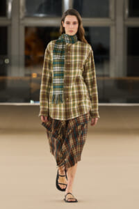Model walks a fashion runway in an oversized plaid shirt, a green plaid scarf, and a fringed plaid skirt, paired with black sandals.