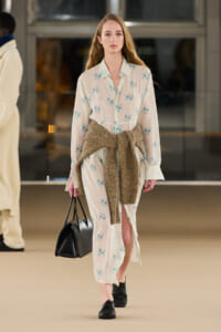 Female model walks a runway in a cream floral shirt-dress with a metallic gold belt, carrying a black handbag and wearing black loafers.