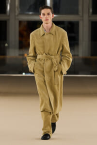 Model walks the runway in a tan corduroy belted jumpsuit, hands in pockets, black shoes visible.