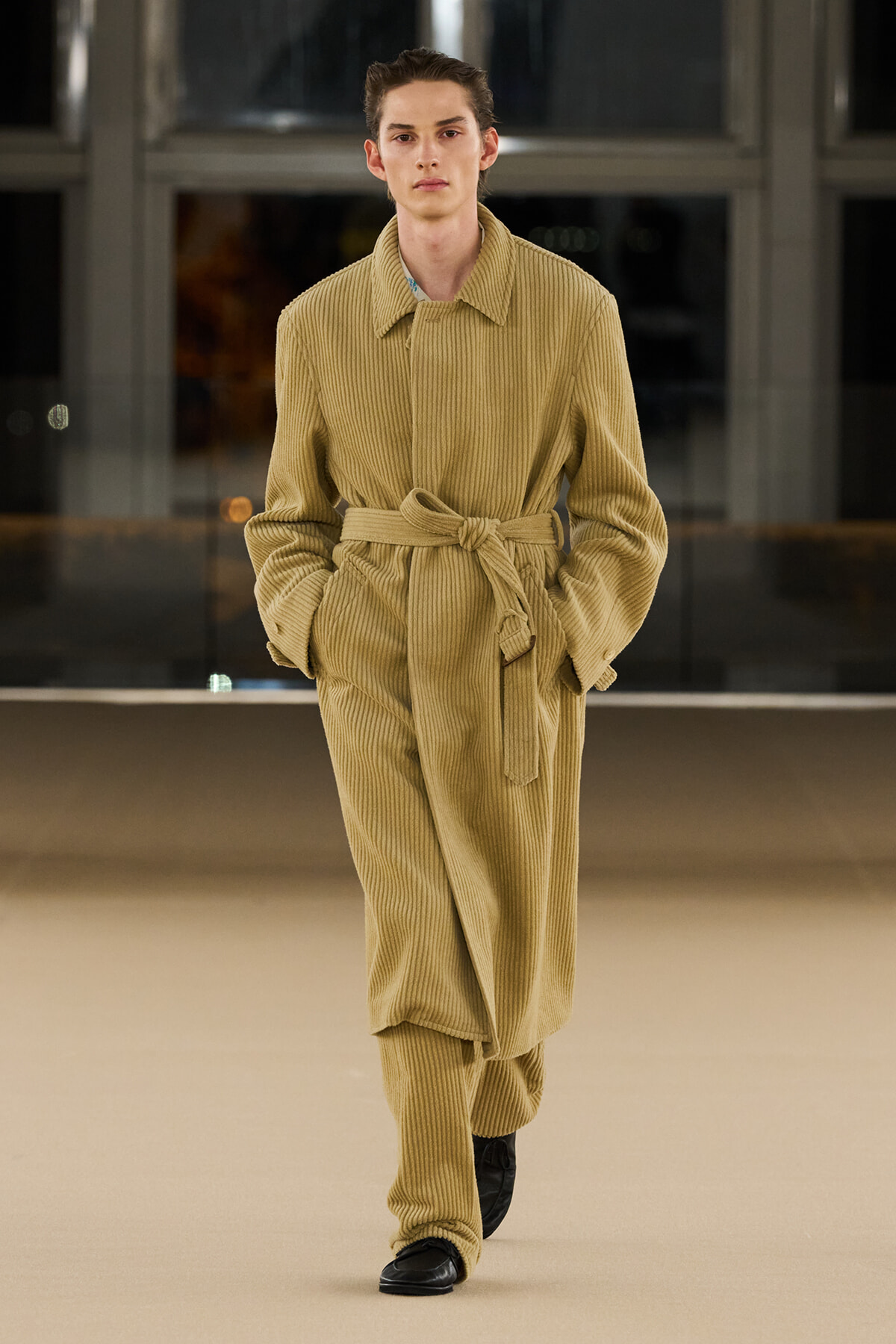 Model walks the runway in a tan corduroy belted jumpsuit, hands in pockets, black shoes visible.