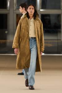 Man with shoulder-length hair wearing a tan long coat, cream sweater, and light-blue jeans walking indoors.