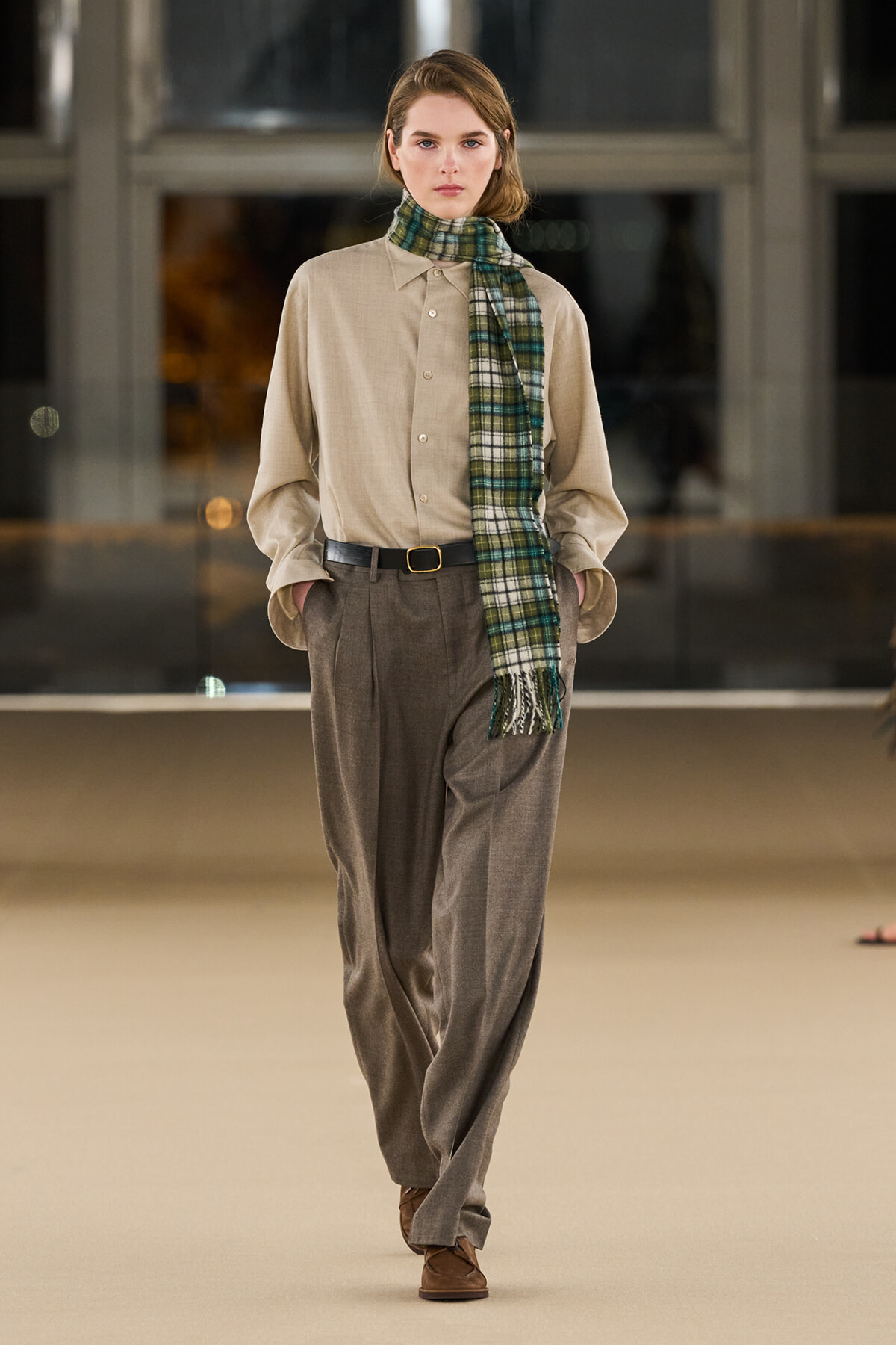 Model walking the runway in a beige button-down shirt, loose gray trousers, brown belt, and a green plaid scarf draped at the neck.