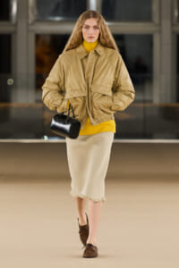 Model in a tan oversized jacket over a mustard yellow turtleneck, beige mid-length skirt with fringe, brown loafers, and a small black handbag.