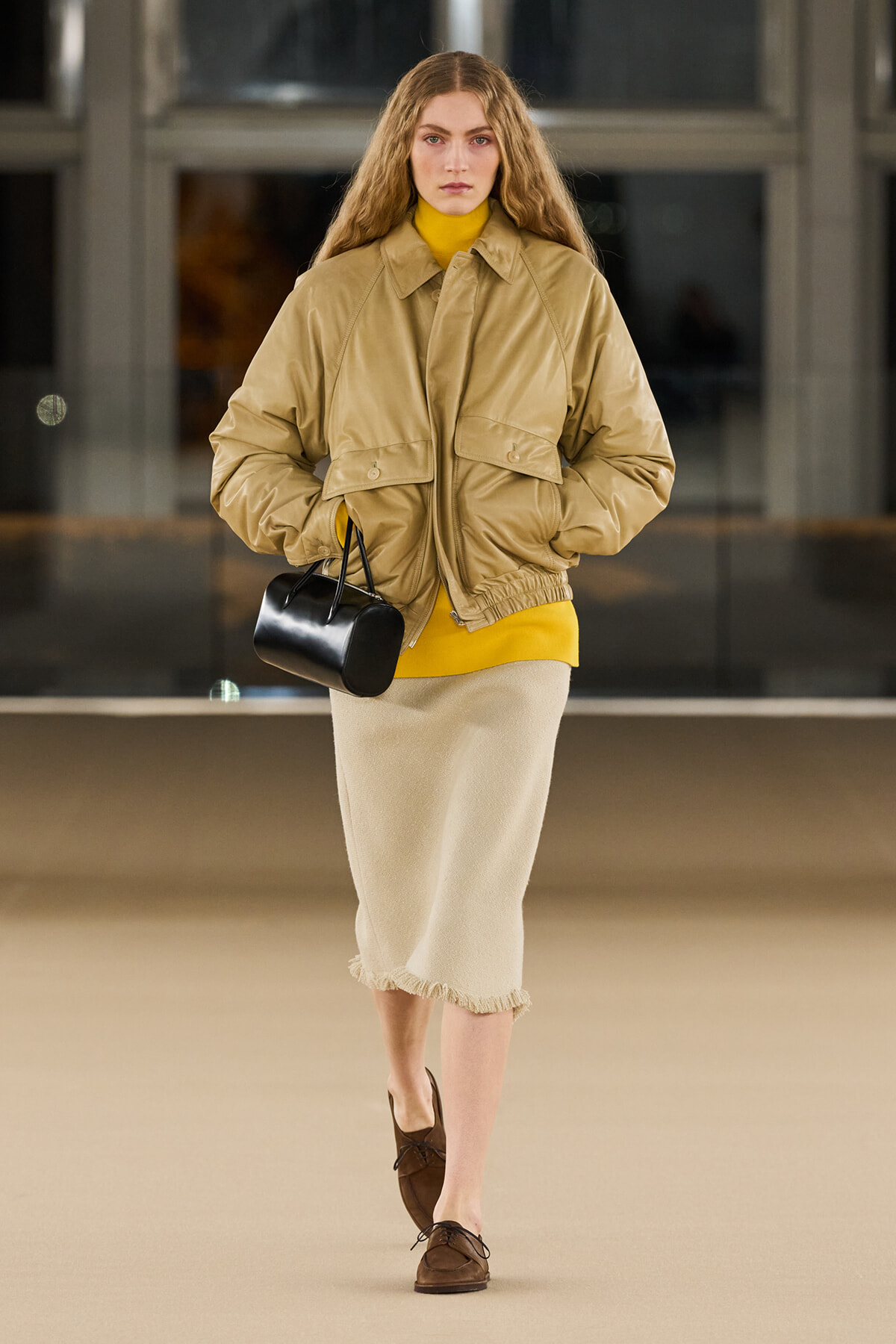 Model in a tan oversized jacket over a mustard yellow turtleneck, beige mid-length skirt with fringe, brown loafers, and a small black handbag.