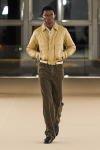 Model walking on a runway in a tan bomber jacket, brown corduroy pants, and a white belt, hands in pockets.
