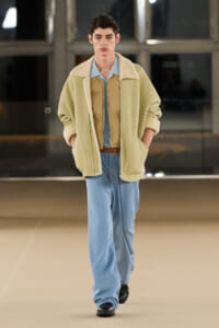 Male model walks a runway in a pale yellow shearling-lined coat, light blue shirt, and loose blue trousers with a brown belt.