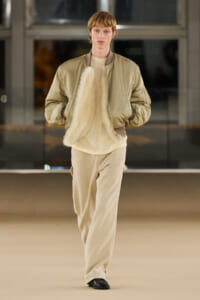 Model walking a fashion runway in a beige bomber jacket, cream sweater, and wide beige pants, holding a fluffy fur scarf.