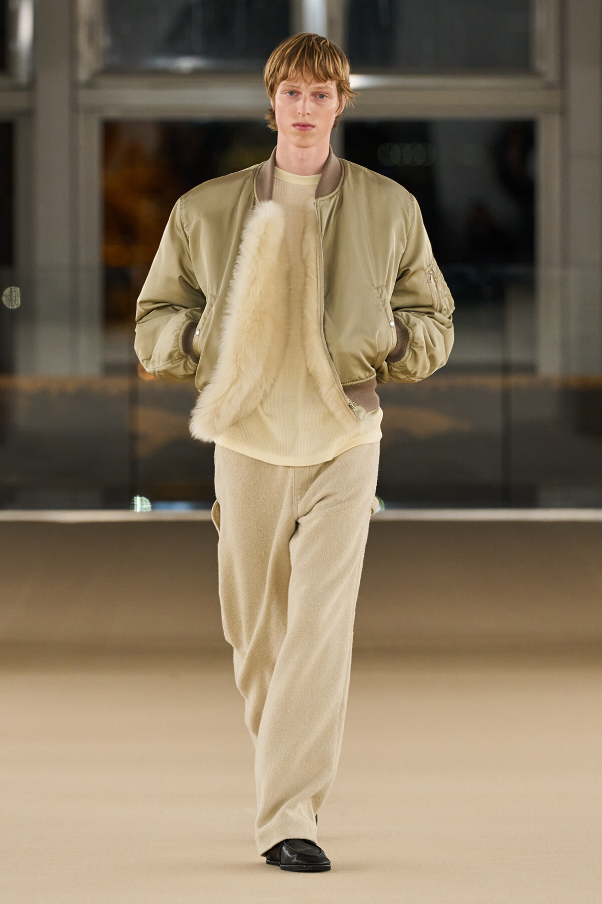 Model walking a fashion runway in a beige bomber jacket, cream sweater, and wide beige pants, holding a fluffy fur scarf.