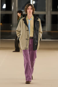 Male model walking a runway in a beige and black oversized jacket, pale yellow shirt, light blue tie, and plum corduroy pants with brown shoes.