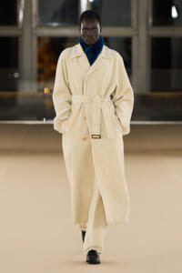 Model walking the runway in a cream corduroy trench with a blue turtleneck, belt tied at the waist.