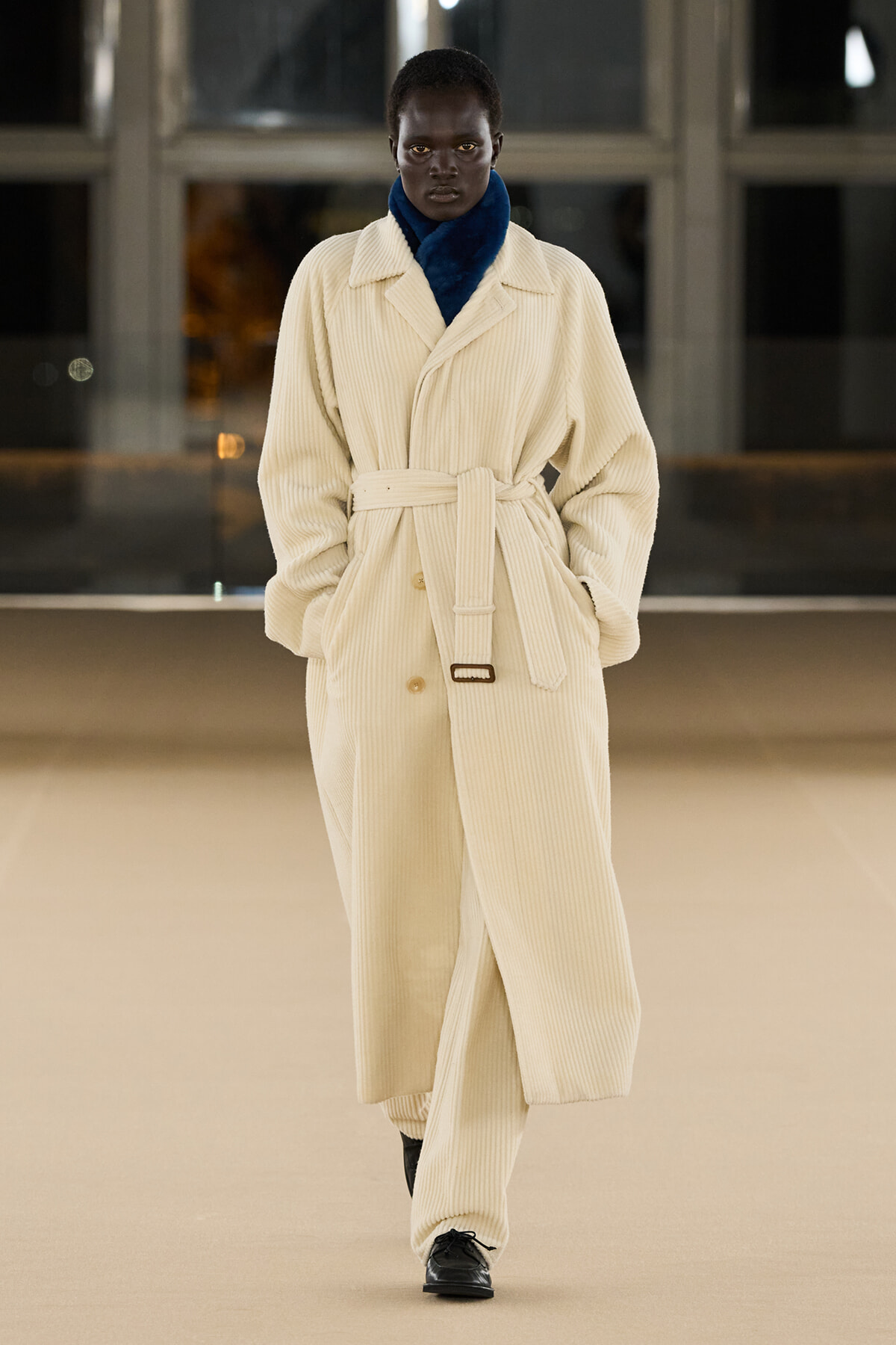 Model walking the runway in a cream corduroy trench with a blue turtleneck, belt tied at the waist.