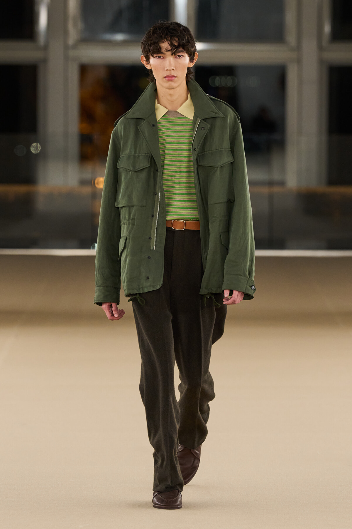 Male model walking a runway in an olive green utility jacket over a striped green shirt, with a brown belt and dark trousers.