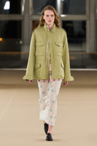 Model walking the runway in an olive green utility jacket over a pink plaid shirt, paired with white floral pants and black loafers.