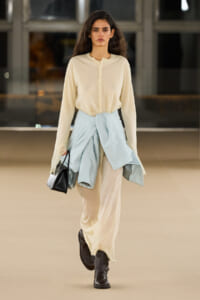 Fashion model walking a runway in a cream blouse, wide beige trousers, with a light blue jacket tied at the waist and black boots, carrying a black handbag.