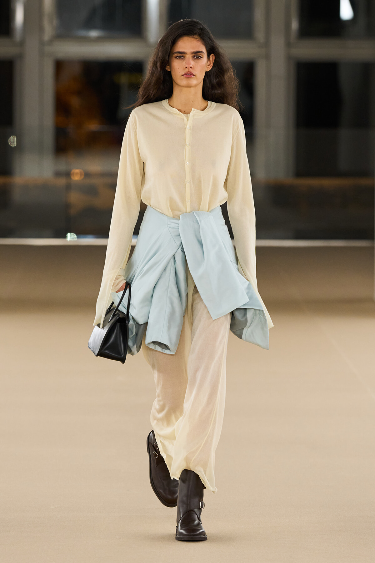 Fashion model walking a runway in a cream blouse, wide beige trousers, with a light blue jacket tied at the waist and black boots, carrying a black handbag.