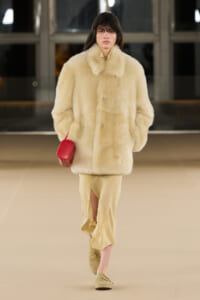 Model on a runway wearing a cream fur coat, beige slitted pants, tan loafers, and a small red handbag.