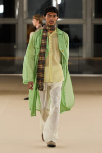 Male model walks a runway in a mint green oversized coat over a pale yellow shirt, white pants, and a plaid scarf.