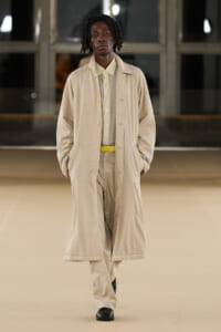 Male model walking a runway in a tan overcoat with matching suit, pale shirt and a yellow belt.