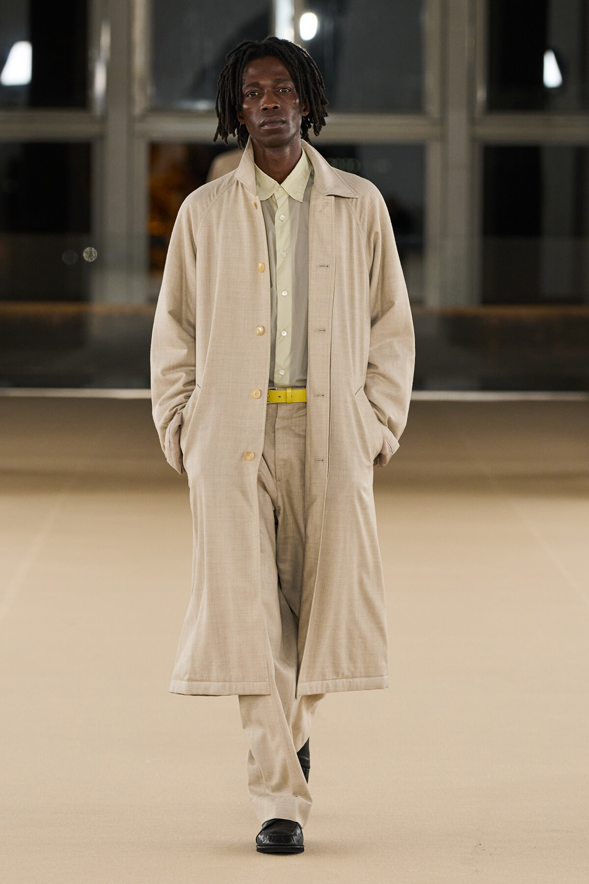 Male model walking a runway in a tan overcoat with matching suit, pale shirt and a yellow belt.