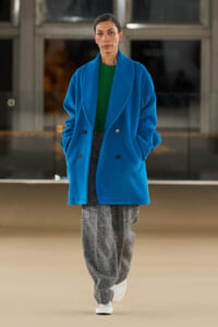 Person standing indoors wearing a bright blue oversized coat over a green sweater and gray wide-leg pants, with white sneakers.