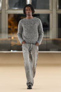 Male model walking on runway in a textured gray sweater and light-washed jeans, hands in pockets, neutral expression.