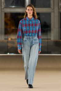 Model walking a fashion runway in a blue and red plaid button-down shirt tucked into high-waisted light-wash jeans, black shoes.