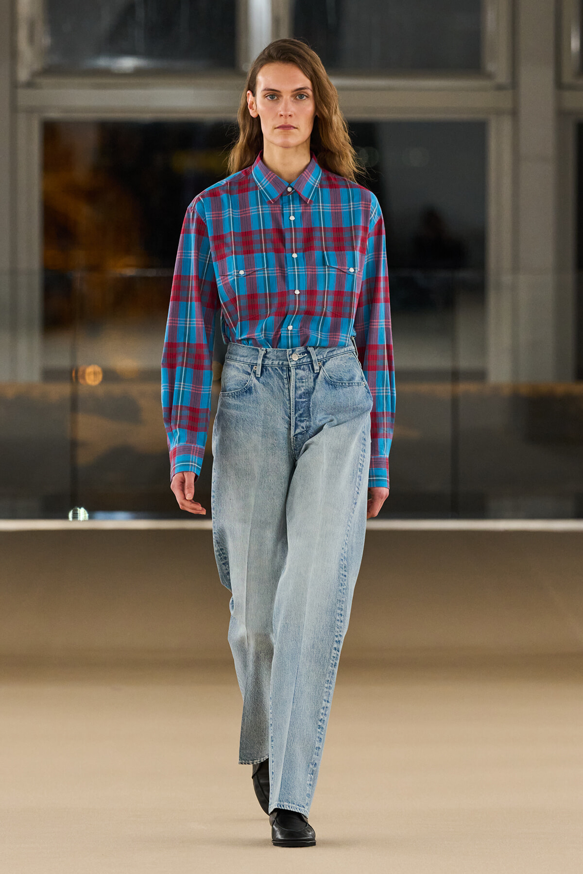 Model walking a fashion runway in a blue and red plaid button-down shirt tucked into high-waisted light-wash jeans, black shoes.