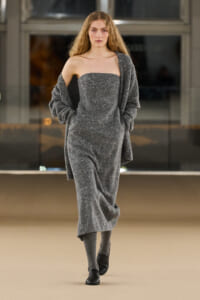 Model walking a fashion runway in a gray, off-shoulder, sparkly jumpsuit with a matching draped cardigan and black shoes.