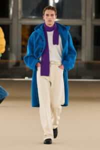 Runway model in a bright blue long coat, white knit shirt, cream trousers, and a purple scarf walking the catwalk with hands in pockets