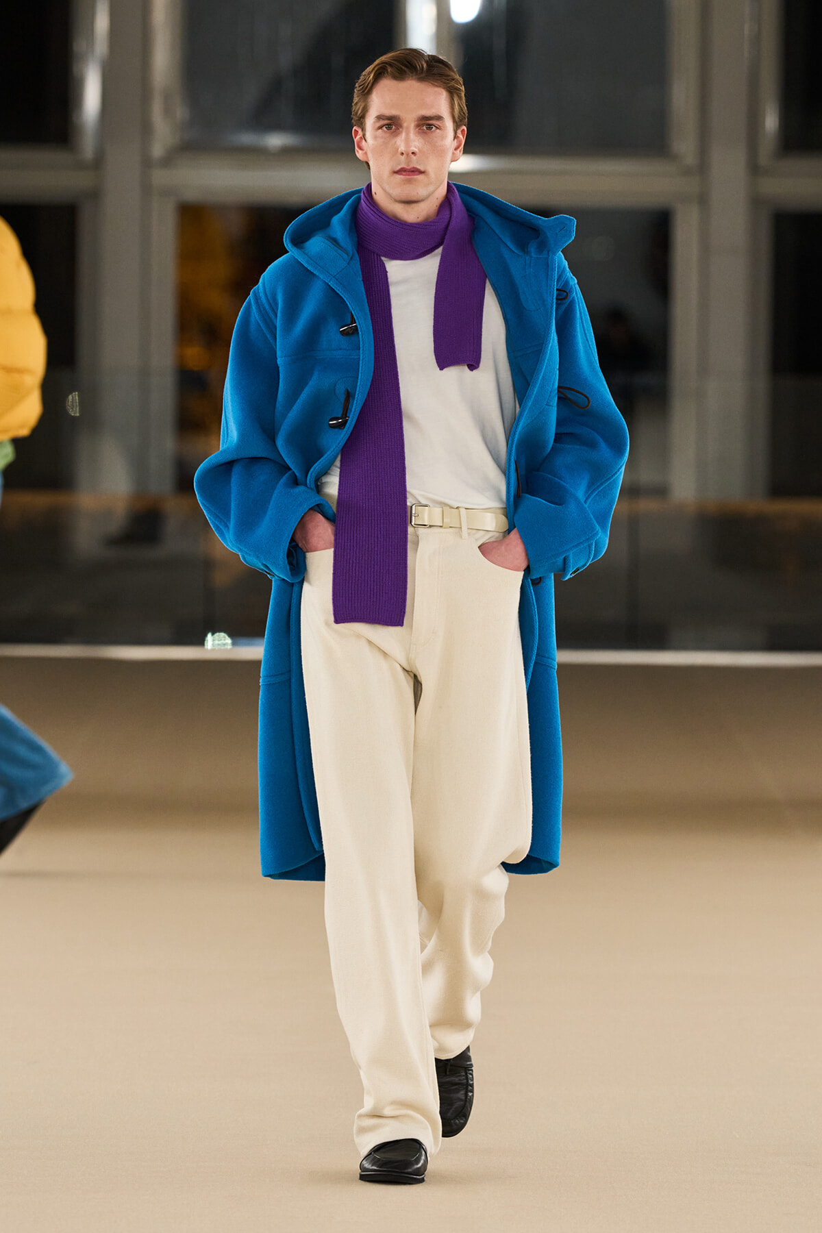 Runway model in a bright blue long coat, white knit shirt, cream trousers, and a purple scarf walking the catwalk with hands in pockets