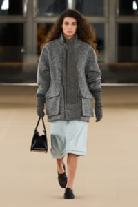 Model walking on a runway in a gray textured oversized coat with matching scarf, light blue knee-length skirt, black loafers, and a small black handbag.