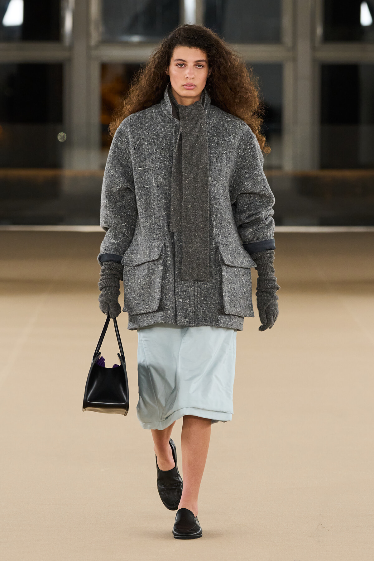 Model walking on a runway in a gray textured oversized coat with matching scarf, light blue knee-length skirt, black loafers, and a small black handbag.