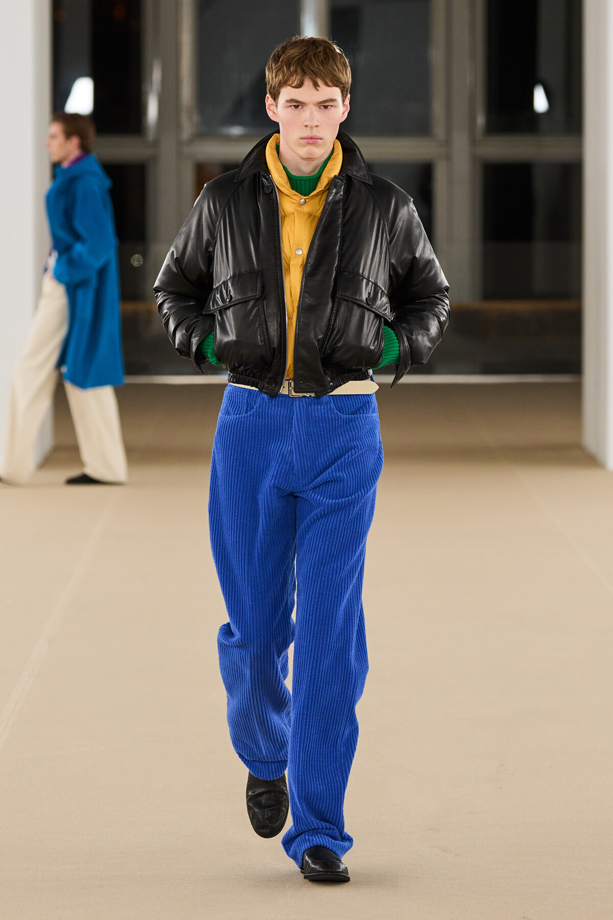 Male model walking a runway in a black leather cropped jacket over a mustard shirt and green knit, paired with bright blue corduroy pants.