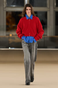 Full-length fashion model walking a runway in a oversized red zip-neck sweater layered over a blue button-down shirt and loose gray jeans, hands in pockets.