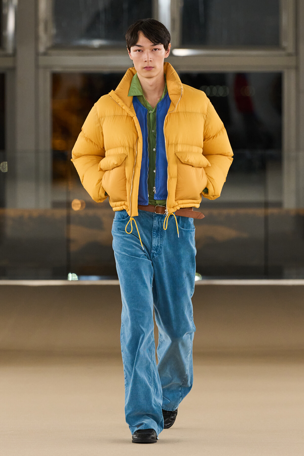 Model wearing a bright yellow puffer jacket over a green shirt and blue jeans, walking toward the camera on a runway-like floor.