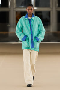 Model walking a runway in an oversized mint-green jacket with blue accents and beige wide-leg pants (full-body shot).