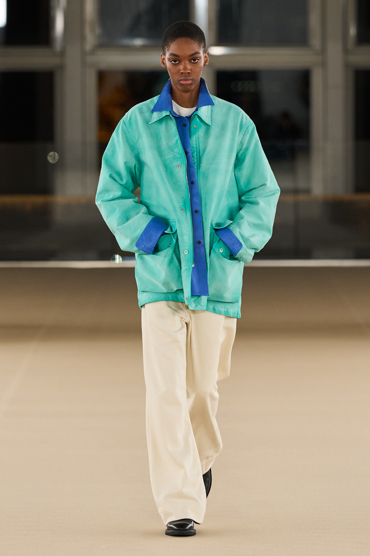 Model walking a runway in an oversized mint-green jacket with blue accents and beige wide-leg pants (full-body shot).
