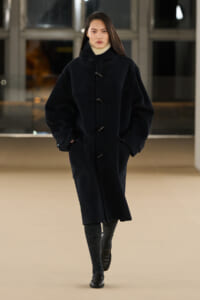 Woman wearing an oversized black duffle coat with toggle closures, hands in pockets, walking indoors in a neutral pose.