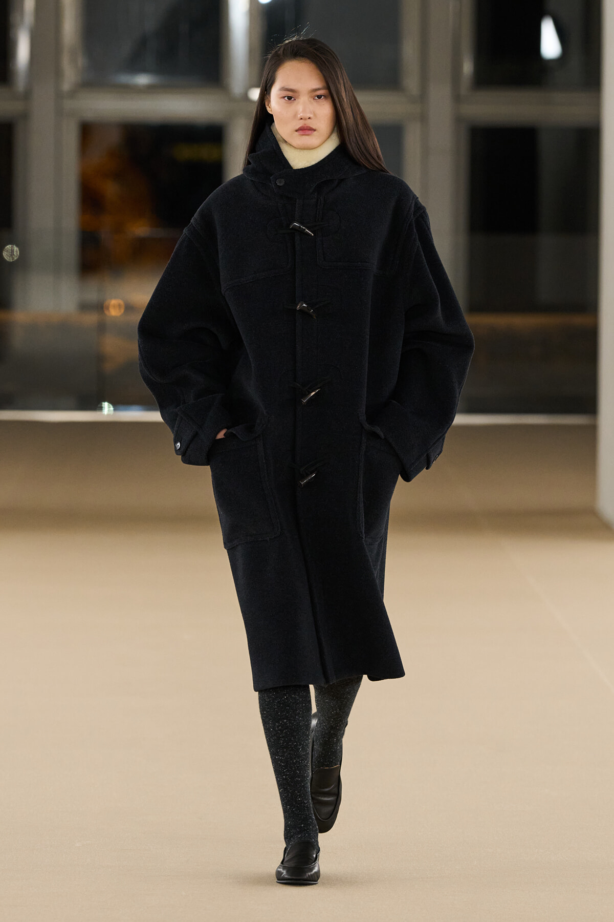 Woman wearing an oversized black duffle coat with toggle closures, hands in pockets, walking indoors in a neutral pose.