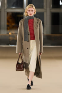 Model walking a runway in a camel wool overcoat over a red striped turtleneck, cream cropped pants, black loafers, gray scarf, and a brown handbag.