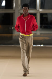 Male model wearing a bright red zip-front jacket over a striped red top, tan wide-leg trousers, yellow belt, and brown shoes walking on a runway in an indoor setting.