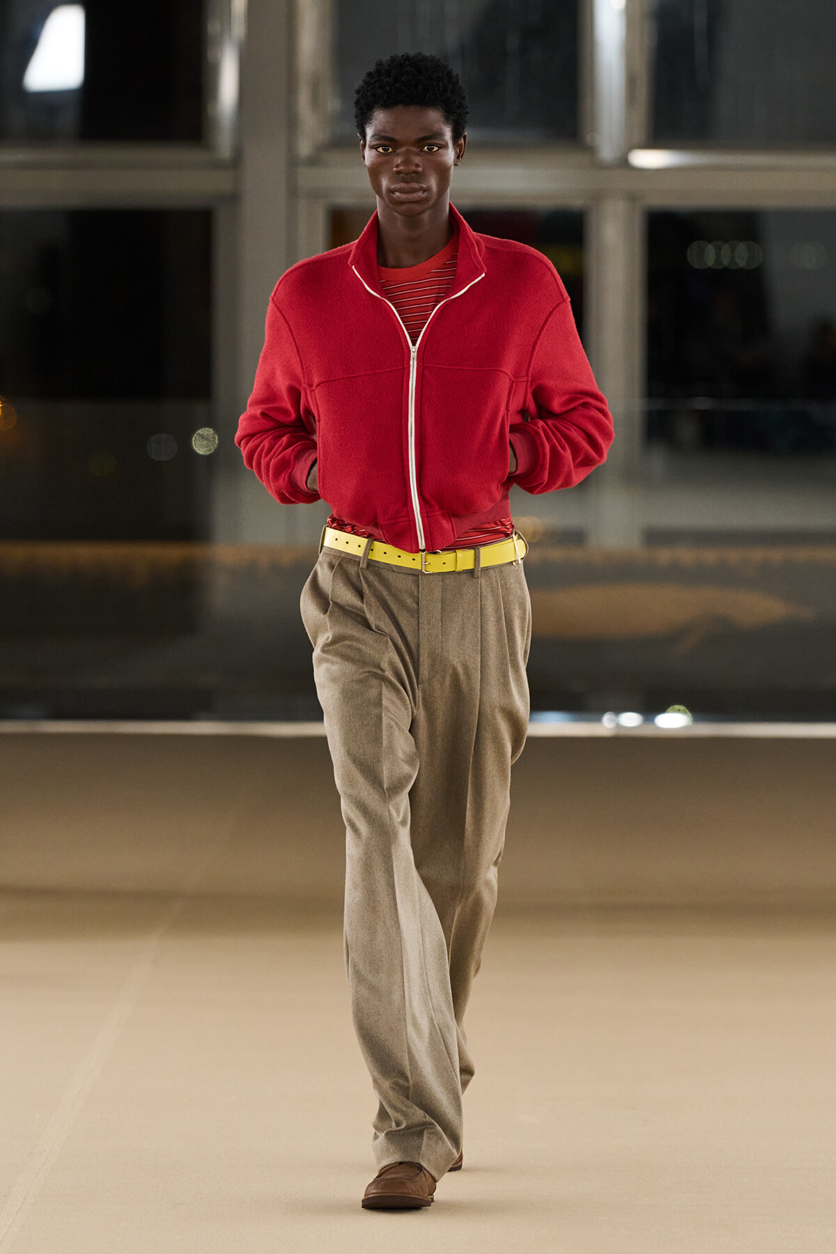 Male model wearing a bright red zip-front jacket over a striped red top, tan wide-leg trousers, yellow belt, and brown shoes walking on a runway in an indoor setting.