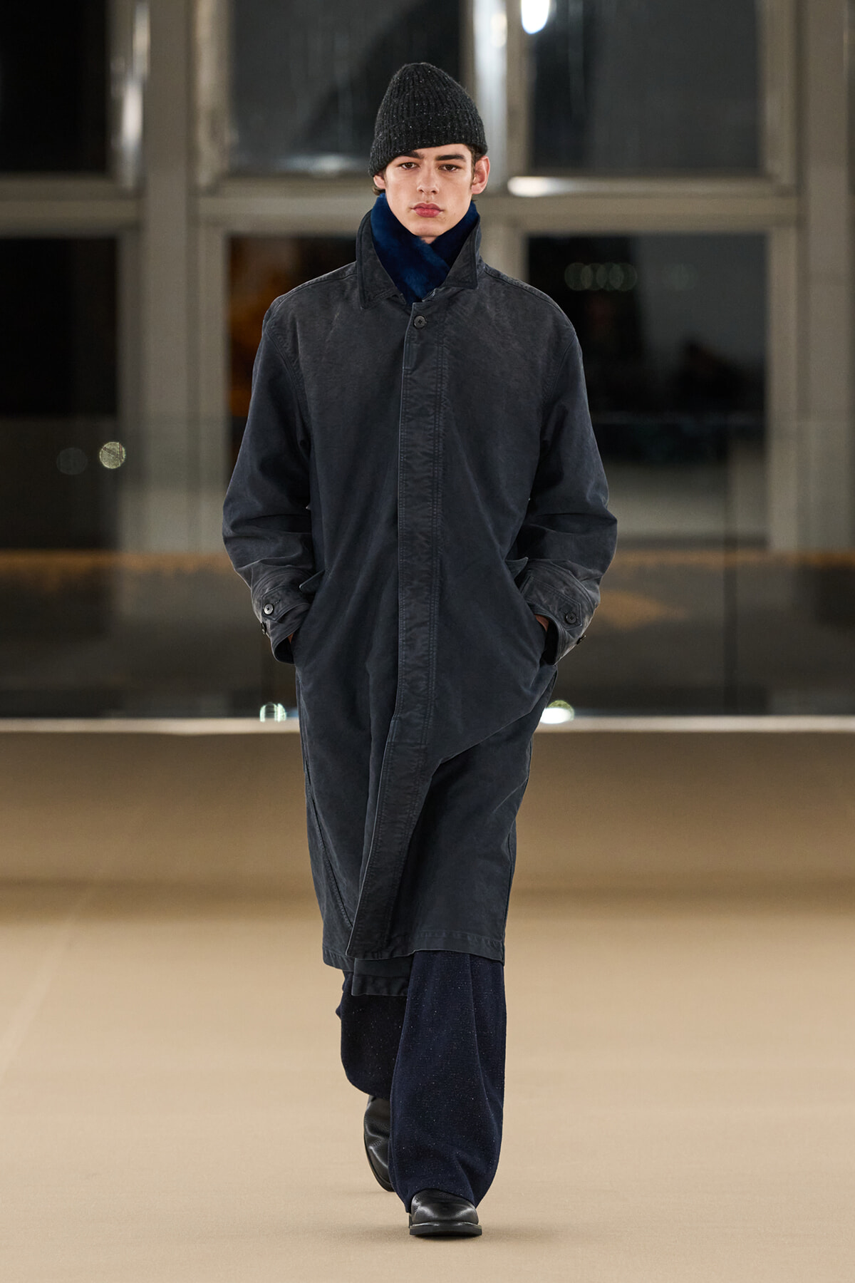 Male model walking a runway in a long dark coat with hands in pockets, wearing a black knit beanie and navy pants.