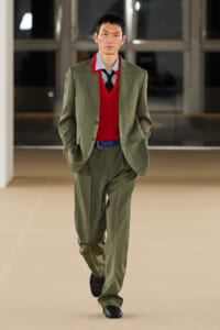 Man in an olive green suit with a red sweater, blue belt, and black shoes walking toward the camera in an indoor hallway.