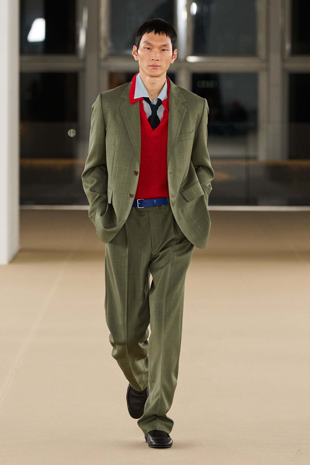 Man in an olive green suit with a red sweater, blue belt, and black shoes walking toward the camera in an indoor hallway.